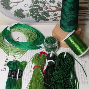 May include: An assortment of green craft supplies, including spools of thread, bundles of cord, embroidery floss, and small glass jars filled with beads. The items vary in shades of green, from light to dark, and are arranged on a white surface.