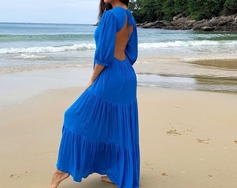May include: A vibrant blue maxi dress with a tiered skirt and open back. The dress has elbow-length sleeves and is worn on a sandy beach with the ocean in the background. The dress is flowing and appears to be made of a lightweight fabric.