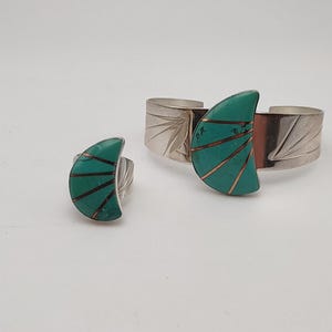 May include: A silver cuff bracelet and a matching ring, both featuring a crescent-shaped inlay of turquoise stone segmented by thin copper lines. The metal bands on both pieces are polished silver with etched fan-like patterns on the sides.