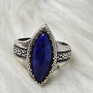 May include: A silver ring featuring a central marquise-shaped deep blue stone. The metal band displays an intricate embossed pattern of small circles and curved lines surrounding the stone and extending along the sides of the ring.