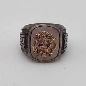 May include: A silver-toned metal ring featuring a central rectangular inlay with a gold-toned eagle emblem. The sides of the ring display embossed lettering that reads USA. The design includes a weathered, dark finish on the metal band.