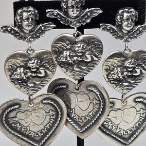 May include: Silver-toned earrings featuring cherub and heart designs. The earrings have three tiers of hearts, with cherub figures at the top and in the middle. The bottom hearts have the text "My Valentine" and a heart pierced by an arrow.