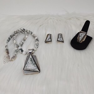 May include: A jewelry set consisting of a necklace, a pair of earrings, and a ring. The necklace features white and black marbled stone beads with a large triangular pendant. The earrings and ring match the pendant, featuring triangular white and black marbled stone inlays set in silver-toned metal frames.