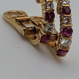 May include: A pair of gold hoop earrings with a hinged clasp. Each earring is adorned with alternating round, deep red gemstones and clear, sparkling stones. The earrings are set against a plain white background.