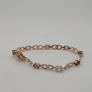 May include: A gold-toned link bracelet featuring a series of oval-shaped multi-colored gemstones interspersed with openwork heart-shaped metal links. The bracelet includes small clear stones set along the metal segments between the larger colored gems.