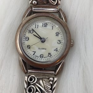 May include: A silver-toned analog wristwatch featuring a round off-white face with black Arabic numerals, three hands, and the text QUARTZ JAPAN MOVT. The watch band is crafted from silver-toned metal with an intricate embossed leaf and scroll pattern.