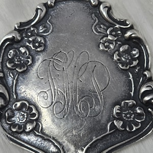 May include: A silver-toned pendant with an ornate, floral-edged design. The pendant features an engraved monogram within a decorative frame, with floral accents. The metal has a slightly aged appearance, suggesting vintage craftsmanship.