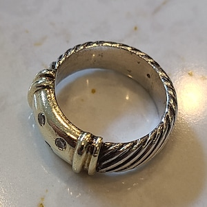 May include: A silver-toned ring with a twisted rope design on the band. The ring features a gold-colored accent with small, clear stones. The ring is a piece of jewelry.