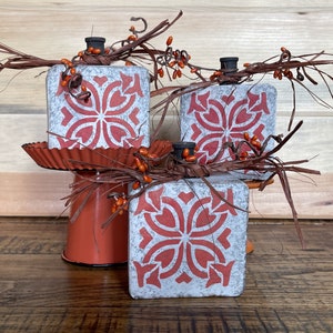 May include: Three decorative, square-shaped pumpkins with a distressed, gray finish and a red floral pattern. Each pumpkin has a black stem, raffia, and orange berry accents. They are arranged on a rustic, orange metal stand, perfect for fall decor.