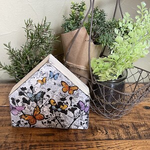 May include: A wooden house-shaped decoration featuring a butterfly design in orange, blue, and purple against a textured gray background. The decor is placed on a wooden surface, with greenery in the background.
