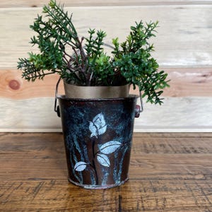 May include: A small, rustic metal planter with a teal and brown distressed finish. The planter has a white floral design and is filled with green artificial foliage.