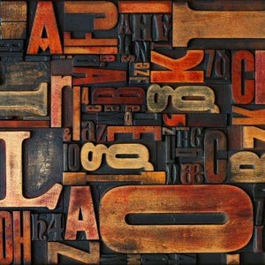 May include: A close-up of a collection of vintage wooden letterpress printing blocks. The blocks are in various sizes and fonts, with warm tones of orange, brown, and gold against a dark background. The arrangement is a mix of letters and numbers.