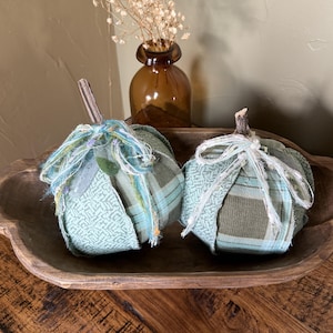 May include: Two handmade fabric pumpkins in a wooden bowl. The pumpkins are made of teal and green patterned fabric, with a frayed ribbon and a twig stem. A brown glass vase with dried flowers is in the background.