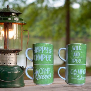 May include: Two sets of stacked green camping mugs with white text. The mugs on the left say "Happy Camper" and "Camp Queen". The mugs on the right say "Wild and Free" and "Campfire Cutie".