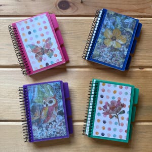May include: Four spiral-bound notebooks with plastic covers and side tabs. The top-left notebook is pink with a butterfly and polka dots. The top-right is blue with a yellow flower. The bottom-left is purple with an owl. The bottom-right is teal with a flower.