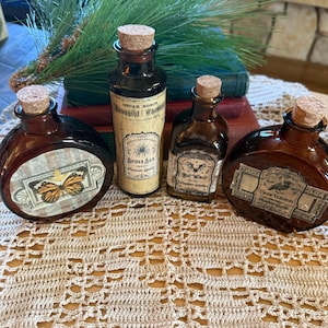 May include: Four amber glass apothecary bottles with cork stoppers. Labels feature illustrations of a butterfly, a spider, a bat, and a crow, with text including Spider Silk, Bat Wings, and Crow Feathers. The bottles are arranged in a group.