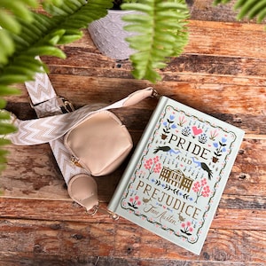 May include: A light blue hardcover book titled "Pride and Prejudice" with floral and bird illustrations. A small, beige crossbody bag with a gold zipper and a patterned strap rests beside the book. The background is a wooden surface.