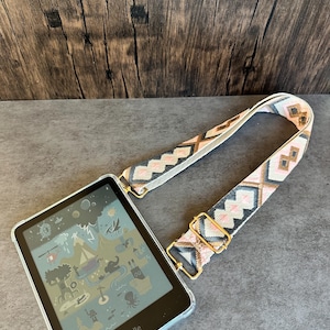 Może przedstawiać: Czytnik e-booków Kindle z przezroczystym etui ochronnym i paskiem we wzory. Pasek ma geometryczny wzór w kolorach różowym, beżowym i brązowym, ze złotymi metalowymi elementami. Ekran Kindle wyświetla kolorową ilustrację.