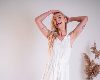 May include: A woman wearing a white sleeveless maxi dress with a tie at the waist. She is smiling and has her hands in her hair.