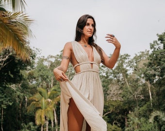 May include: A woman wearing a light beige maxi dress with a plunging neckline and cutouts at the sides. She is standing by a swimming pool with lush greenery in the background.