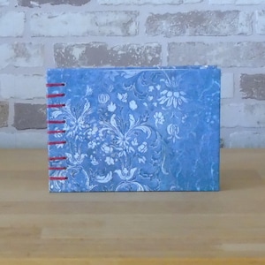 Può includere: Un piccolo diario rettangolare con una copertina blu a motivi floreali. La copertina presenta un motivo floreale bianco ornato. Il dorso è cucito a mano con filo rosso. Il diario è appoggiato su una superficie di legno marrone chiaro.