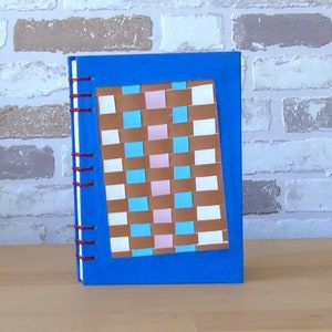 Op de afbeelding: Een blauw notitieboek met een bruin, roze, wit en blauw geruit patroon op de voorkant. Het notitieboek heeft een rode spiraalbinding.