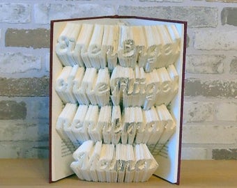 May include: A book with the words "She believed she could, so she did" folded into a 3D sculpture. The book is open and the pages are folded to create the words. The book is on a wooden surface.