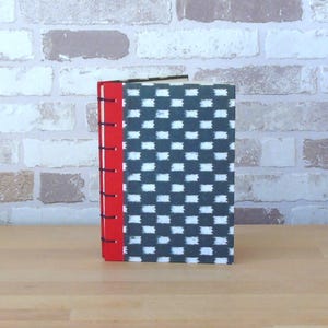 Op de afbeelding: Twee handgemaakte notitieboeken met een rode rug en zwarte stiksels. De covers hebben een zwart-wit geometrisch patroon. De notitieboeken staan op een houten oppervlak tegen een bakstenen muur.