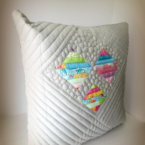 K&ouml;nnte beinhalten: Ein quadratisches, hellgraues, gestepptes Kissen mit drei bunten rautenf&ouml;rmigen Patchwork-Designs. Der Patchwork verwendet verschiedene gemusterte Stoffe in Blau-, Rosa-, Gr&uuml;n- und Gelbt&ouml;nen. Das Kissen hat eine strukturierte, gesteppte Oberfl&auml;che.