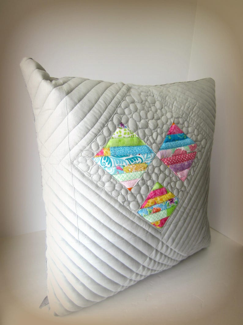 K&ouml;nnte beinhalten: Ein quadratisches, hellgraues, gestepptes Kissen mit drei bunten rautenf&ouml;rmigen Patchwork-Designs. Der Patchwork verwendet verschiedene gemusterte Stoffe in Blau-, Rosa-, Gr&uuml;n- und Gelbt&ouml;nen. Das Kissen hat eine strukturierte, gesteppte Oberfl&auml;che.