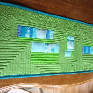 May include: A green quilted table runner with a teal border. The runner features a geometric design with blue and white fabric squares and rectangles.
