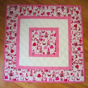 May include: A square, quilted table topper with a Valentine's Day theme. The design features a white center with pink borders and a repeating pattern of gnomes, hearts, and strawberries in shades of red and pink. The outer border is a solid pink.