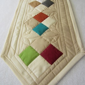 May include: A beige quilted table runner with a diamond pattern of colorful fabric squares. The squares are in shades of red, green, blue, orange, brown, and white.