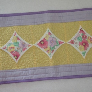 May include: A rectangular quilted table runner with a yellow center and a lavender border. The center features diamond-shaped floral patterns in pink, red, and yellow. The runner is approximately 12 inches wide and 36 inches long.