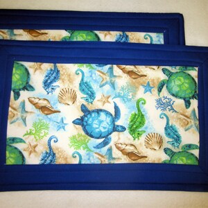 May include: Two blue and white quilted placemats with a sea turtle, starfish, and seashell design. The placemats have a dark blue border.