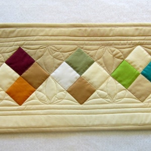 May include: A beige quilted table runner with a row of colorful diamond-shaped fabric patches. The patches are in shades of green, white, brown, orange, teal, and burgundy.