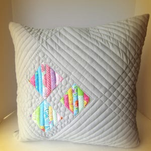 K&ouml;nnte beinhalten: Ein quadratisches, graues, gestepptes Kissen mit einem Rautenmuster. Zwei farbenfrohe rautenf&ouml;rmige Patches zeigen vertikale Streifen aus gemustertem Stoff in Rosa, Blau, Gr&uuml;n und Gelb. Ein dekoratives Wohnaccessoire.