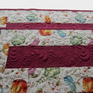 May include: A quilted table runner with a fall theme. The runner is made of a cream-colored fabric with a print of pumpkins, gourds, and leaves. The runner has a burgundy border and a burgundy center panel with a quilted design. The text "Leaves are falling, Autumn is calling" is printed on the runner.