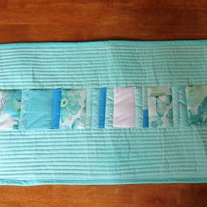 May include: A turquoise table runner with a row of patchwork squares in various shades of blue and white with floral and butterfly patterns.