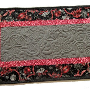 May include: A gray quilted table runner with a red and white border. The border features a Christmas-themed print with ornaments, bows, and bells. The runner is approximately 16 inches wide and 30 inches long.
