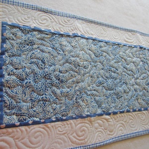 May include: A blue and white patterned table runner with a white background and a blue and white gingham border. The runner is quilted with a swirl pattern.