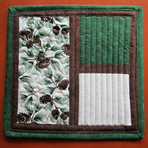 May include: A green quilted placemat with a brown border and a white center featuring a pine cone pattern. The placemat is divided into three sections, with the center section featuring a white fabric with vertical quilting.