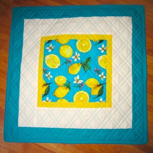 May include: Square quilted placemat with a vibrant lemon pattern on a turquoise background. The placemat features a white border and a turquoise outer edge, creating a cheerful and colorful design.