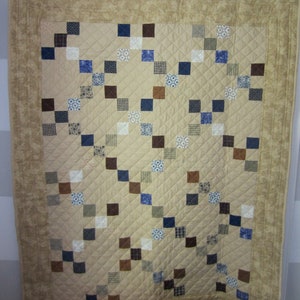 May include: A beige quilt with a brown and blue patchwork design. The quilt is made up of small squares of fabric, each with a different pattern. The quilt is edged with a brown border.