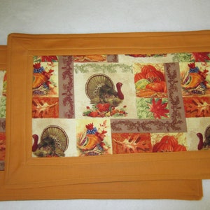 May include: Two orange fabric placemats with a Thanksgiving theme. The placemats feature a patchwork design with images of turkeys, pumpkins, and fall leaves.