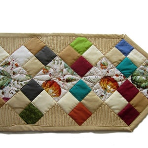 May include: A quilted table runner with a diamond pattern in various colors, including green, red, blue, and brown. The runner features a central design of pumpkins and leaves with the text "It's Fall" in the center.