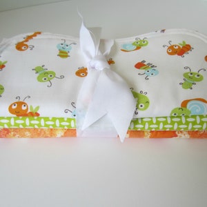 Puede incluir: Un conjunto de tres baberos para bebé con fondo blanco y un patrón de insectos coloridos. Los baberos están atados con una cinta blanca.