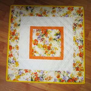 May include: Square quilted placemat with a floral design. The center features a square of orange and yellow flowers, framed by a white quilted square. The outer border has a floral pattern with a yellow edge.