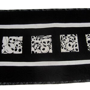 May include: Black and white table runner with a repeating pattern of Jack Skellington's face from the Nightmare Before Christmas. The runner has a white border and a black background.