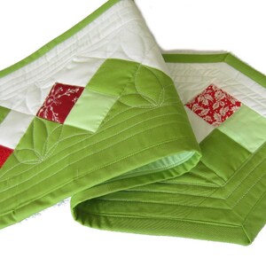 May include: A green quilted table runner with a white background and red and green patchwork squares. The runner has a decorative stitch pattern around the edges.